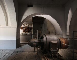 Sala macchine della centrale idroelettrica dell’Ex Lanificio Carotti: generatore storico in primo piano e quadro elettrico con strumenti sotto le volte in mattoni intonacati