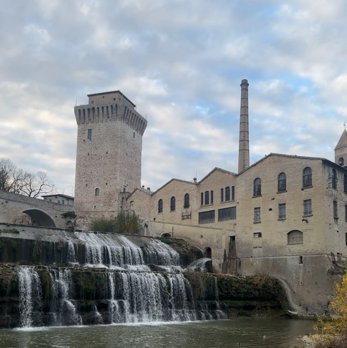 Cortile esterno dell'Ex Lanificio Carotti sulla cascata del Metauro, con la torre medievale e la ciminiera in mattoni, spazio all'aperto per pause e attività di team building