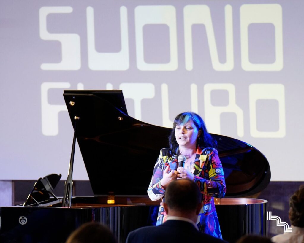 Presentazione di Suono Futuro all'Ex Lanificio Carotti: relatrice al microfono davanti a un pianoforte a coda, con la scritta Suono Futuro proiettata sulla parete e il pubblico in primo piano