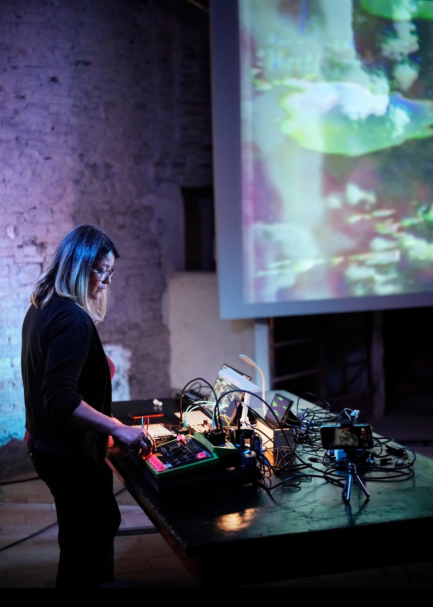 Una performer al banco degli strumenti elettronici, illuminata dai riflessi colorati sulla parete