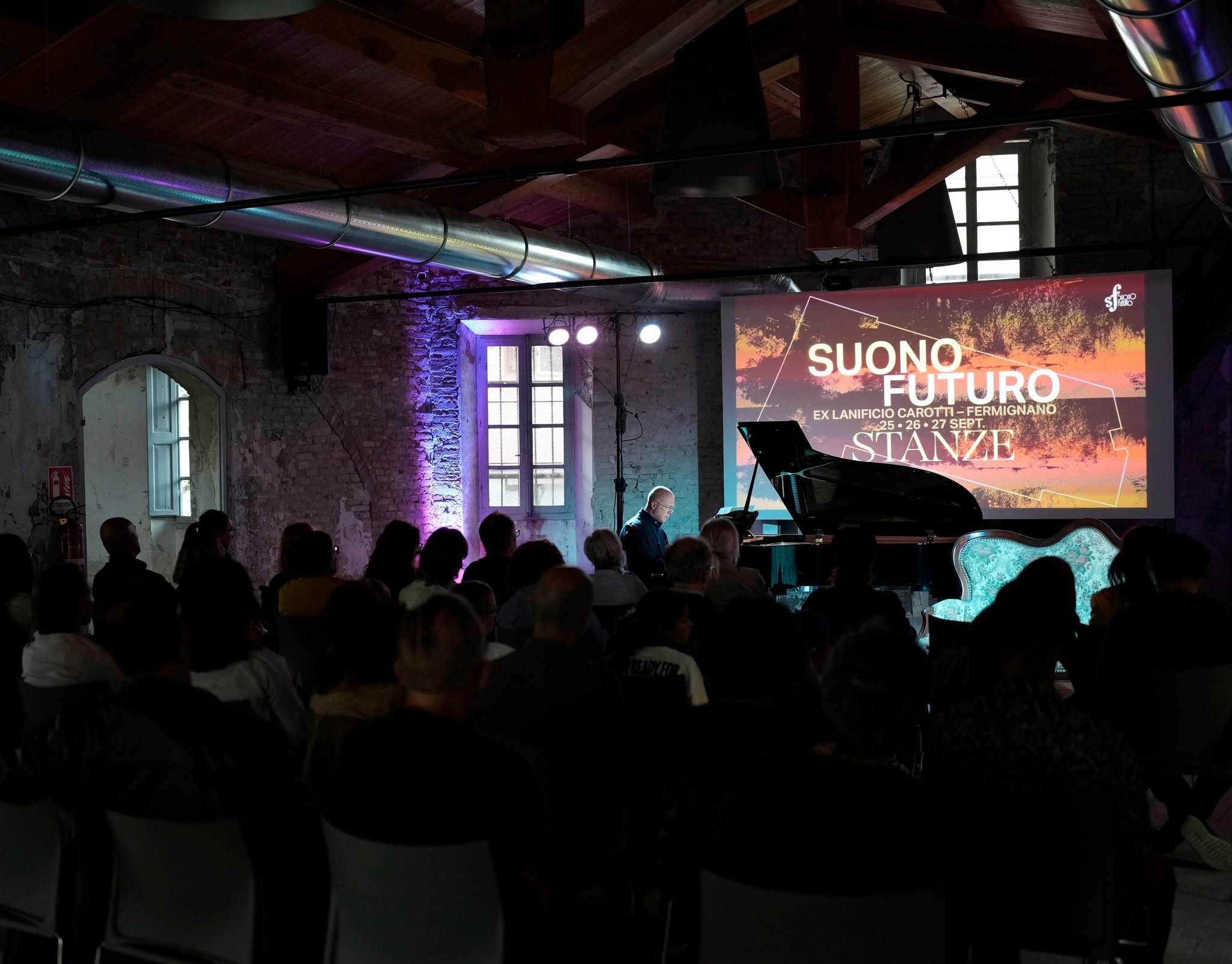 Il pubblico assiste al recital pianistico di chiusura, con il divano vintage e la scritta Stanze sullo sfondo